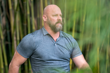 Portrait of a macho man with beard and bald head. Green leafs blurry in foreground