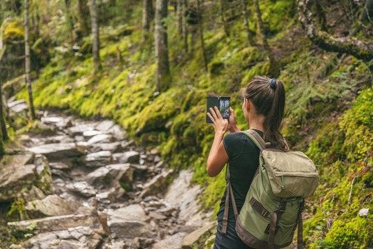 Hiker girl tourist taking picture with mobile phone of trail in nature forest hiking in Quebec outdoors fall autumn season, Canada travel lifestyle. Woman walking with backpack.