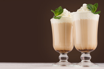 Latte coffee in two elegant transparent glass with whipped cream, green fresh mint in modern dark brown kitchen interior, copy space.