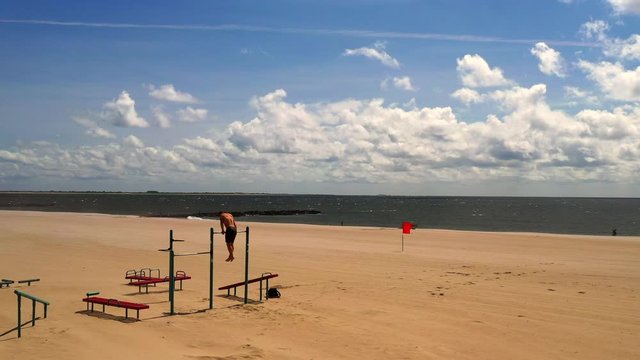 drone camera steady shot, as a young man exercises on a pull up bar on Coney Island beach in Brooklyn, NY at a workout station with the sandy beach, the Atlantic Ocean, the blue sky & horizon in view