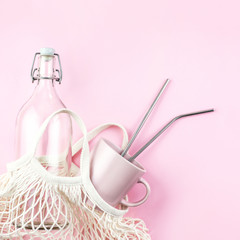 Mesh bag with reusable glass water bottle and cup on pink background.