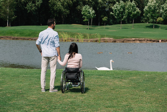 Back view of young man holding hands with disabled woman while looking at pond in park - Powered by Adobe