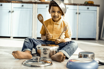 Boy sitting on the kitchen floor and playing on a saucepan while wearing a colander on the head.