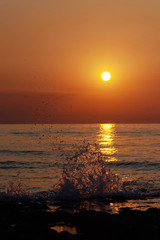 Romantic sunset by a stony seashore. The sun sets over the horizon. The sun beams reflecting in the calm sea waters. Stony shore is washed by the gentle waves. The sky is turning yellow and orange.