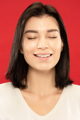 Caucasian young woman's close up portrait on red studio background. Beautiful female model in white shirt with well-kept skin. Concept of human emotions, facial expression. Looks calm and dreamful.