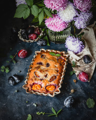 baked plum pie in ceramic mold among fresh plums and flowers on blue background