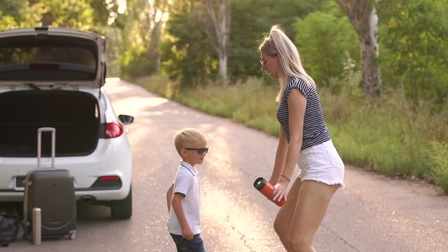 Cheerful young mother funny dancing with her son on the road in the woods near the car during the summer trip. Slow motion. Family having fun while traveling by car.
