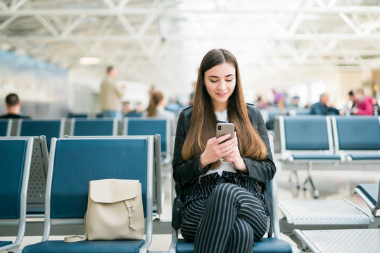 Airport Young female passenger on smart phone at gate waiting in terminal while waiting for her flight. Air travel concept with young casual woman sitting with hand luggage suitcase.