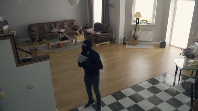 The thief in black clothes and balaclava walking in large living room. The guy stealing tablet and noticing security camera. Concept of crime, offense against the law. Top view, camera moving closer