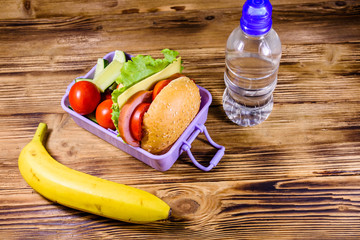 Banana, bottle of water and lunch box with hamburger, cucumbers and tomatoes on wooden table