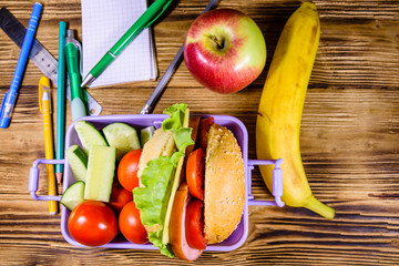 Ripe apple, banana, different stationeries and lunch box with hamburger, cucumbers and tomatoes on wooden table. Top view