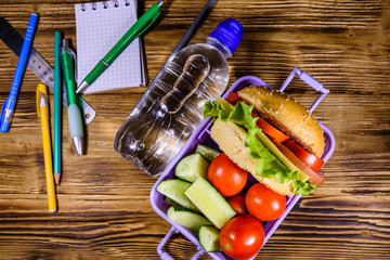 Bottle of water, different stationeries and lunch box with sandwiches, cucumbers and tomatoes on wooden table. Top view