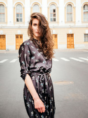 portrait of a young beautiful brown haired woman with long wavy hair in a brown dress  in the city against a yellow building,