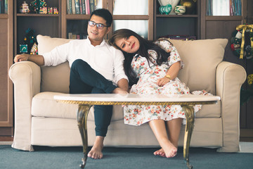Asian male and female couples, in which men wear white shirts and women wear floral dresses, smiling while sitting on a sofa in a warm home.