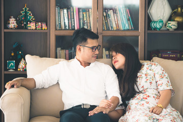 Asian male and female couples, in which men wear white shirts and women wear floral dresses, smiling while sitting on a sofa in a warm home.