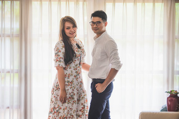 Asian male and female couples in which men wear white shirts and women wear floral dresses while standing by the window with white curtains in a warm home.