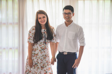 Asian male and female couples in which men wear white shirts and women wear floral dresses while standing by the window with white curtains in a warm home.