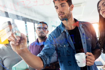 Successful young man writing on office glass board during the meeting with colleagues in the coworking space.