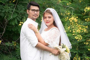 Portrait of young Asian wedding couple smiling and holding flowers in park.
