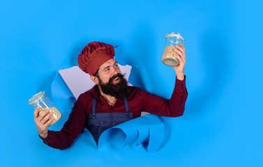 Diet, organic food, vegetarian, groats concept. Bearded chef holds in hands jars with groats. Male chef in hat and apron looking through paper hole. Healthy diet concept. Cook holds jars with cereal.