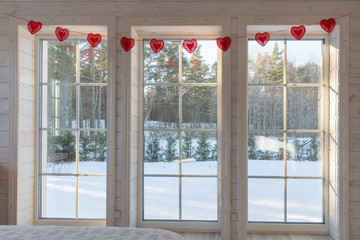 A large white bedroom window in wooden house decorated with romantic garland of red hearts on Valentine's Day.