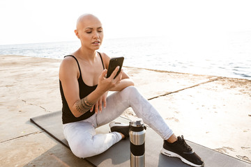 Fototapeta na wymiar Image of serious bald woman typing on cellphone while sitting on mat