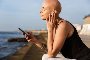 Image of relaxed bald woman using cellphone and earphone while sitting