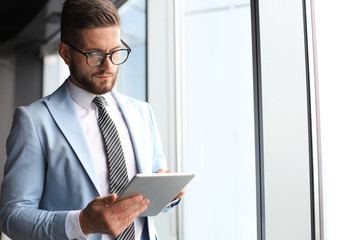 Modern business man in formalwear using digital tablet while standing near window in the office