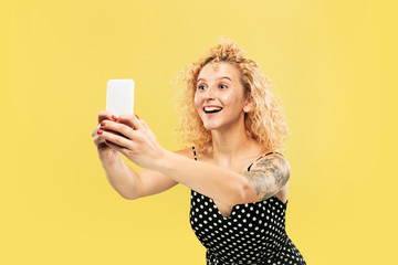 Caucasian young woman's half-length portrait on yellow studio background. Beautiful female model in black dress. Concept of human emotions, facial expression. Taking selfie or her own vlog, smiling.