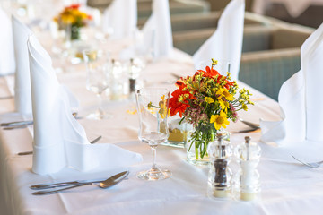 A restaurant. Festive table setting. Bouquet of orange and yellow flowers, glasses, plates, cutlery, white napkins and candles on a white tablecloth