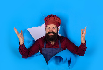 Bearded man in chef uniform looking through paper hole. Professional approach to business. Professional happy man chef. Cooking, profession and business. Male chef, cook or baker in hat and apron.