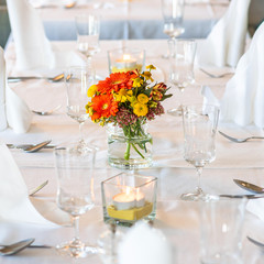 A restaurant. Festive table setting. Bouquet of orange and yellow flowers, glasses, plates, cutlery, white napkins and candles on a white tablecloth