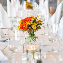 A restaurant. Festive table setting. Bouquet of orange and yellow flowers, glasses, plates, cutlery, white napkins and candles on a white tablecloth
