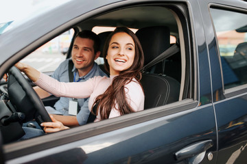 Male auto instructor takes exam in young woman. Cheerful attractive female driver posing at driver's place. Look from car. Hold hands on sheering wheel.