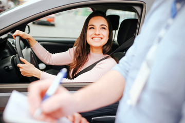 Fototapeta na wymiar Male auto instructor takes exam in young woman. Cheerful attractive model look at guy and smile. Holding hands on steering wheel. Instructor writing mark on paper.