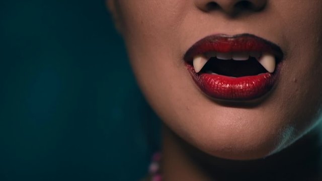 Sexy Vampire Woman's red bloody lips close-up. Vampire girl licking fangs with tongue. Fashion Glamour Halloween art design. Close up of female vampire mouth, teeth. On black background 4K UHD video