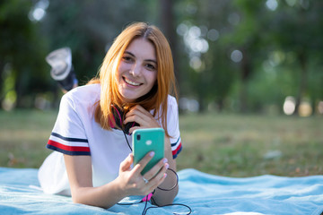 Pretty smiling teenage girl with red hair using sellphone outdoors in park.
