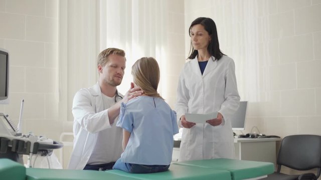 Pediatrician checking tonsils of little girl and listening to her lung sounds with stethoscope, female nurse standing nearby holding medical history of lab test results