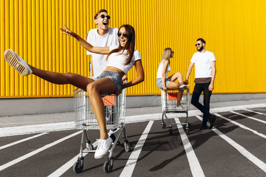 A group of young people in white t-shirts, fun ride on carts near the store, supermarket, friends have fun in Sunny weather outdoors