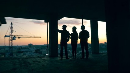 Backlit architects talking in a building under construction during sunset.