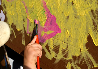 A kid's hand with a paint brush painting on cardboard house with yellow and pink color outside on sunny day