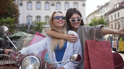 Two happy winning female friends with shop packages sitting on the cool scooter and laughing on the background of city buildings