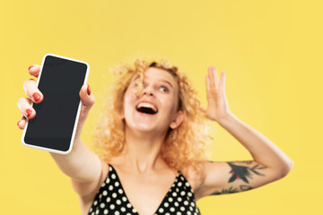 Caucasian young woman's half-length portrait on yellow studio background. Beautiful female model in black dress. Concept of human emotions, facial expression. Showing phone's screen, looks crazy happy