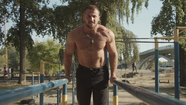 Shirtless determined athletic fit man with well trained body doing triceps dips on parallel bars in outdoor gym. Muscular bodybuilder working out arms, training triceps on dips horizontal bars.