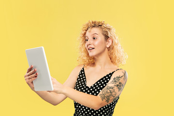 Caucasian young woman's half-length portrait on yellow studio background. Beautiful female model in black dress. Concept of human emotions, facial expression. Using tablet for vlog or selfie.