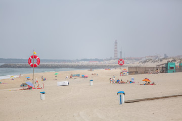View at the beach with people taking sunbath on beach, lifeguard lifebuoys and a beach bar, lighthouse as background