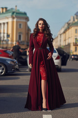 Naklejka premium Young beautiful stunning woman in red luxury evening dress with flying hem walking city street on a sunny evening. Elegant lady with makeup and wavy brunette hair. Full length fashion portrait