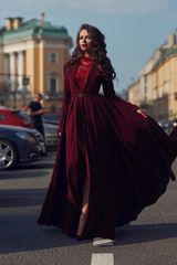 Naklejka premium Young beautiful stunning woman in red luxury evening dress with flying hem walking city street on a sunny evening. Elegant lady with makeup and wavy brunette hair. Full length fashion portrait