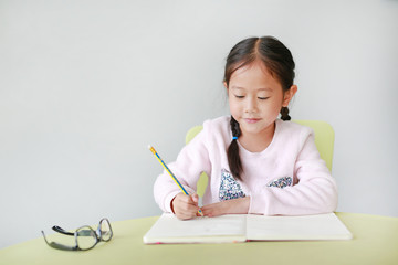 Happy little Asian child girl write in a book or notebook with pencil sitting on kid chair and table against white background.