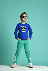 Kid boy in cool blue aviator sunglasses, blue shirt with print is standing with his hands in pockets of his green pants